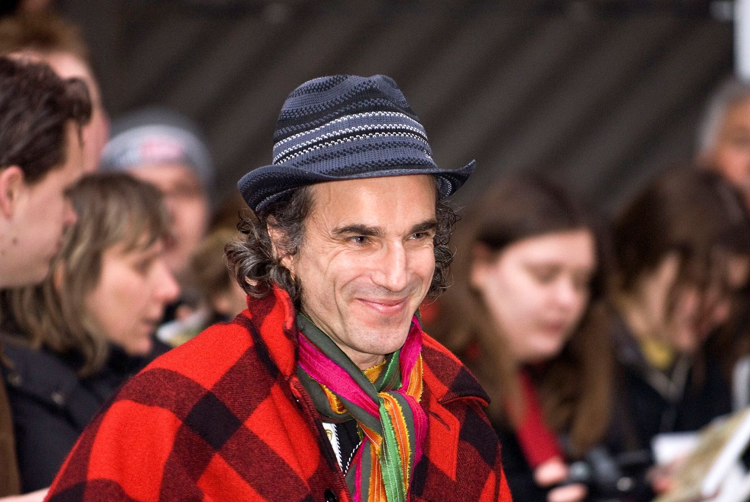 Daniel Day-Lewis en manteau à carreaux rouge et chapeau, souriant à la Berlinale 2008, foule floue en arrière-plan.