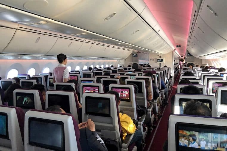 Cabine d’avion en classe économique presque pleine, passagers installés avec écrans dossiers allumés et éclairage d’ambiance au plafond.