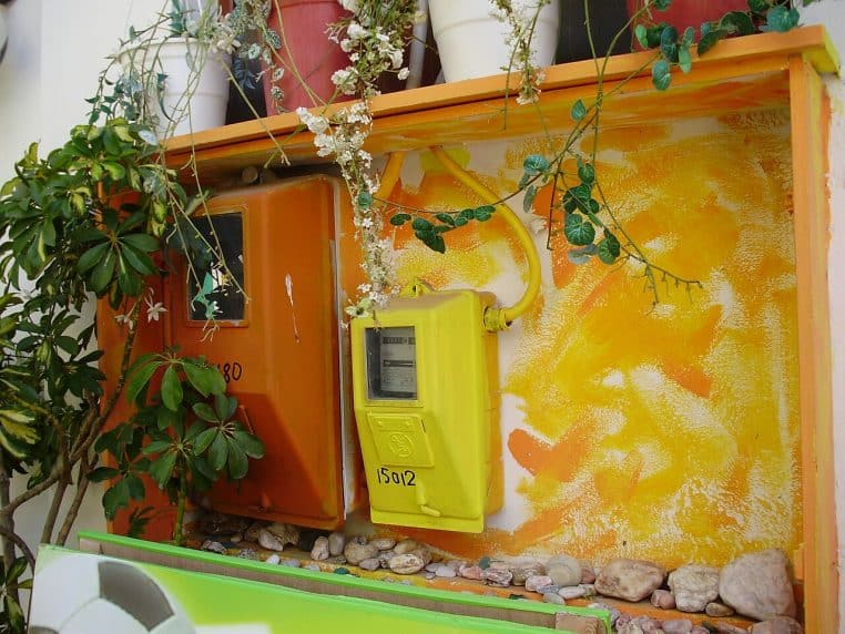 Compteur électrique installé sur un mur en crépi gris, cadré large, coffret et câbles visibles en extérieur