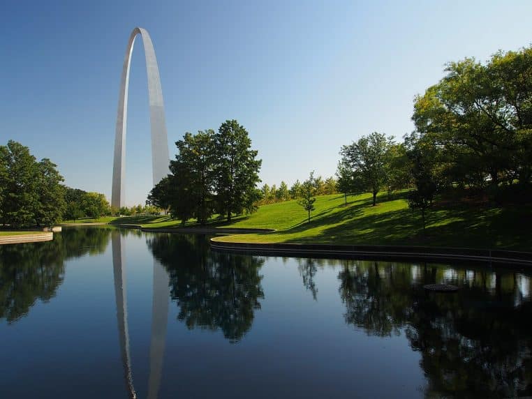 La Gateway Arch se reflète dans un bassin du parc ; ciel clair et arbres au premier plan, format paysage.