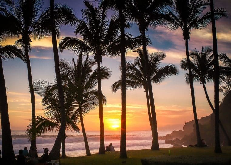 Plage de Grand Anse à la Réunion au crépuscule, houle régulière et falaises verdoyantes sous un ciel orangé.