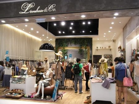 Intérieur de boutique de vêtements avec portants et présentoirs, perspective large en format horizontal, ambiance lumineuse et contemporaine.