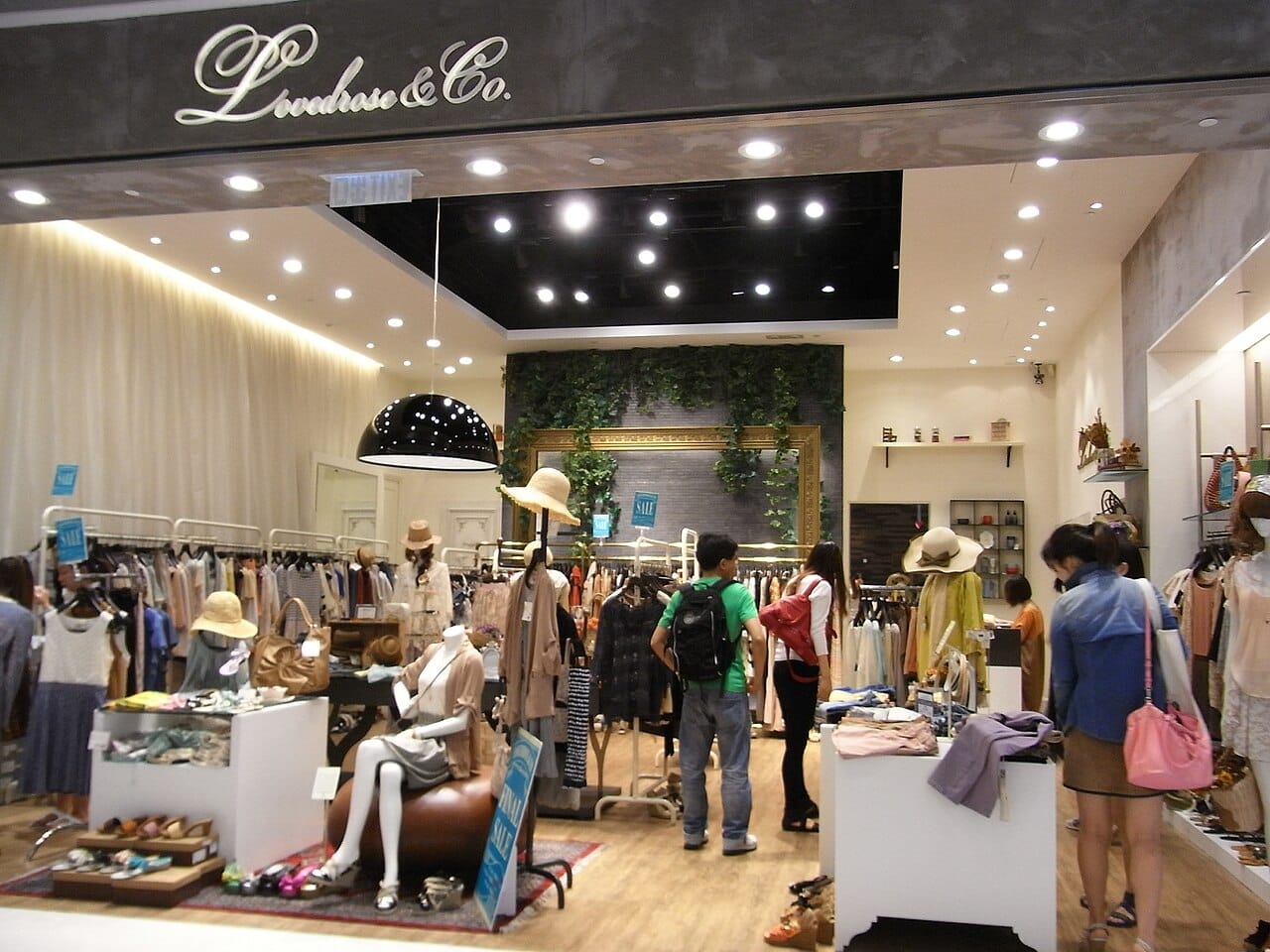 Intérieur de boutique de vêtements avec portants et présentoirs, perspective large en format horizontal, ambiance lumineuse et contemporaine.