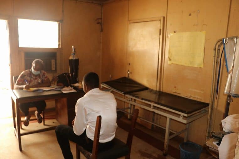 Professionnelle de santé conseillant un patient en face à face dans un bureau, documents et lit d’examen à côté.