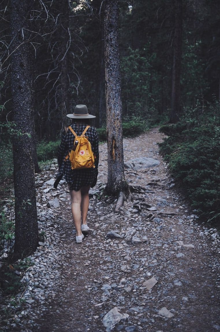 Randonneuse avec sac sur un sentier caillouteux en forêt, dos à l’objectif, progression tranquille vers la clairière.