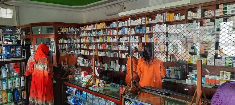Vue large d’une pharmacie avec de multiples boîtes de médicaments alignées sur des étagères lumineuses, illustrant le coût des soins en 2026.