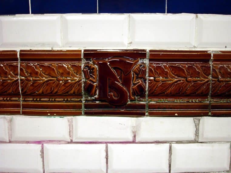 Gros plan horizontal des carreaux à motifs Nord-Sud avec encadrement brun et lettrage, typique des stations historiques.