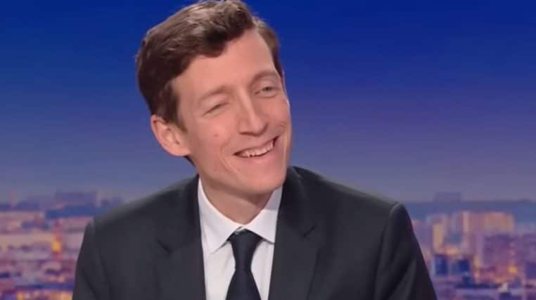 Plan rapproché d’un homme en costume sombre souriant sur un plateau télévisé, avec un fond urbain flou au crépuscule.