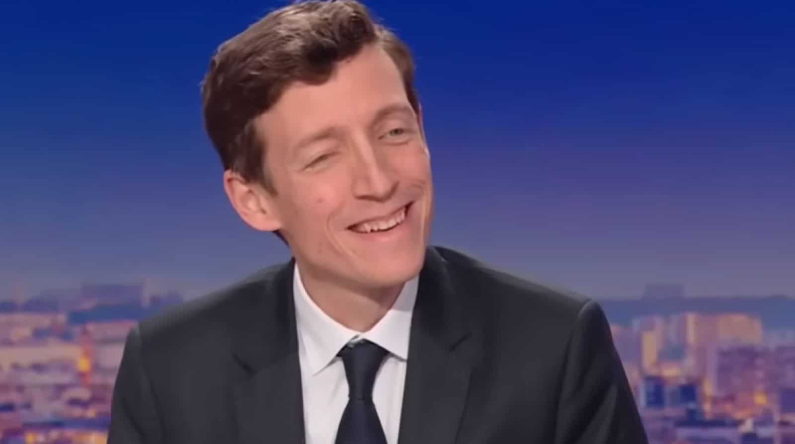 Plan rapproché d’un homme en costume sombre souriant sur un plateau télévisé, avec un fond urbain flou au crépuscule.