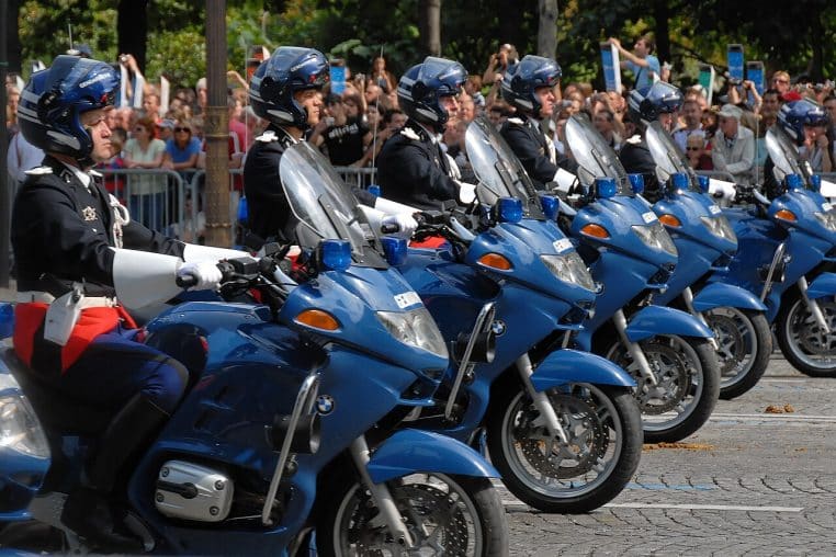 Deux motos de la gendarmerie départementale en formation lors d’un défilé, vues de trois-quarts, circulation coupée et public en arrière-plan.
