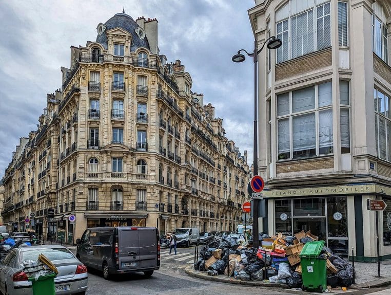 Rangées de sacs et conteneurs débordant sur un trottoir parisien, illustrant la pression sur la collecte et le traitement des déchets.