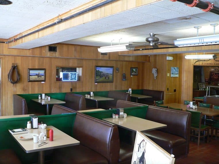 Rangées de banquettes brun-vert dans un restaurant à boiseries, tables dressées simplement, ambiance rétro.