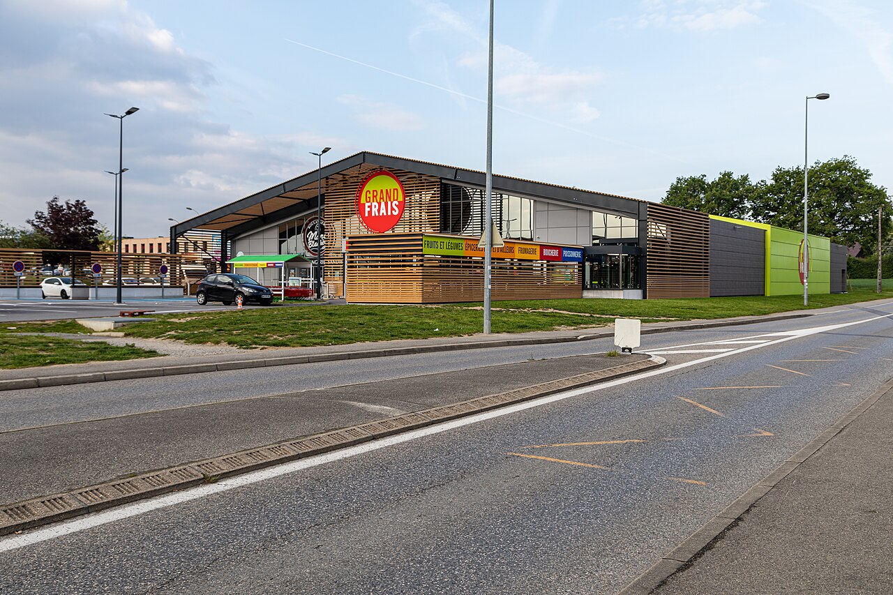 Entrée d’un Grand Frais à Ferney-Voltaire, ciel couvert, parking au premier plan.