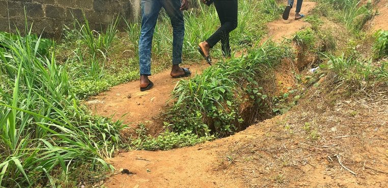 Chemin de terre bordé d’herbes où marchent des personnes, cadrage sur les jambes et le sentier.