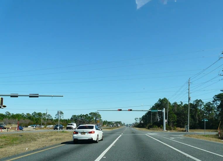 Feu tricolore suspendu au-dessus d’une large chaussée à un carrefour routier, vue horizontale en plein jour, circulation fluide aux abords.