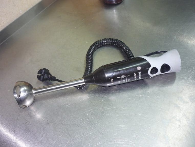 Mixeur plongeant Bosch et gobelet doseur posés sur un plan de travail, câble visible, vue horizontale en lumière intérieure.