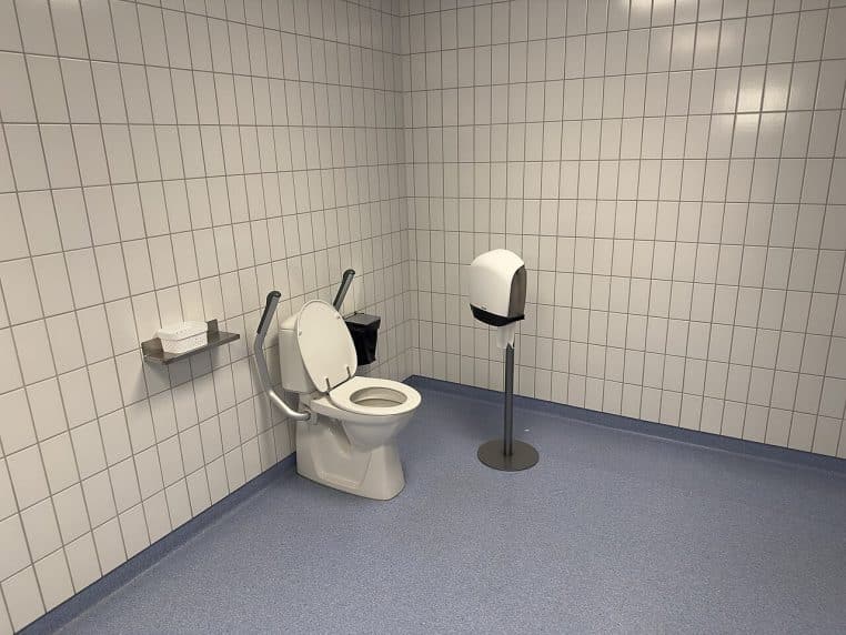 Vue d’un sanitaire accessible très propre, avec cuvette, barres d’appui et accessoires, éclairage blanc et carrelage mural.