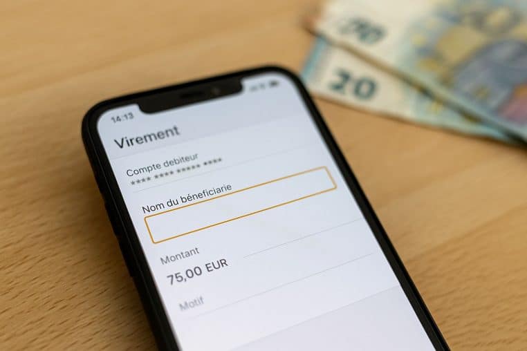 Écran de virement bancaire sur smartphone avec le champ « Nom du bénéficiaire » à renseigner et des billets d’euros flous en arrière-plan.