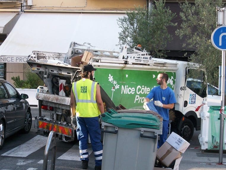 : Deux agents de propreté assurent la collecte des déchets à Nice, avec camion et bacs, en tenue haute visibilité.