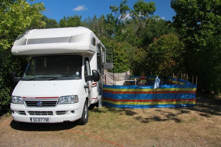 Camping-car blanc stationné dans un champ, ciel nuageux et montagnes lointaines.