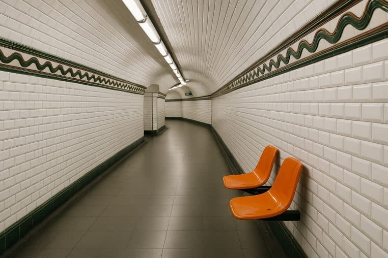 Couloir du métro parisien vide, murs en carrelage blanc biseauté avec frise verte et brune, deux sièges orange, éclairage néon.