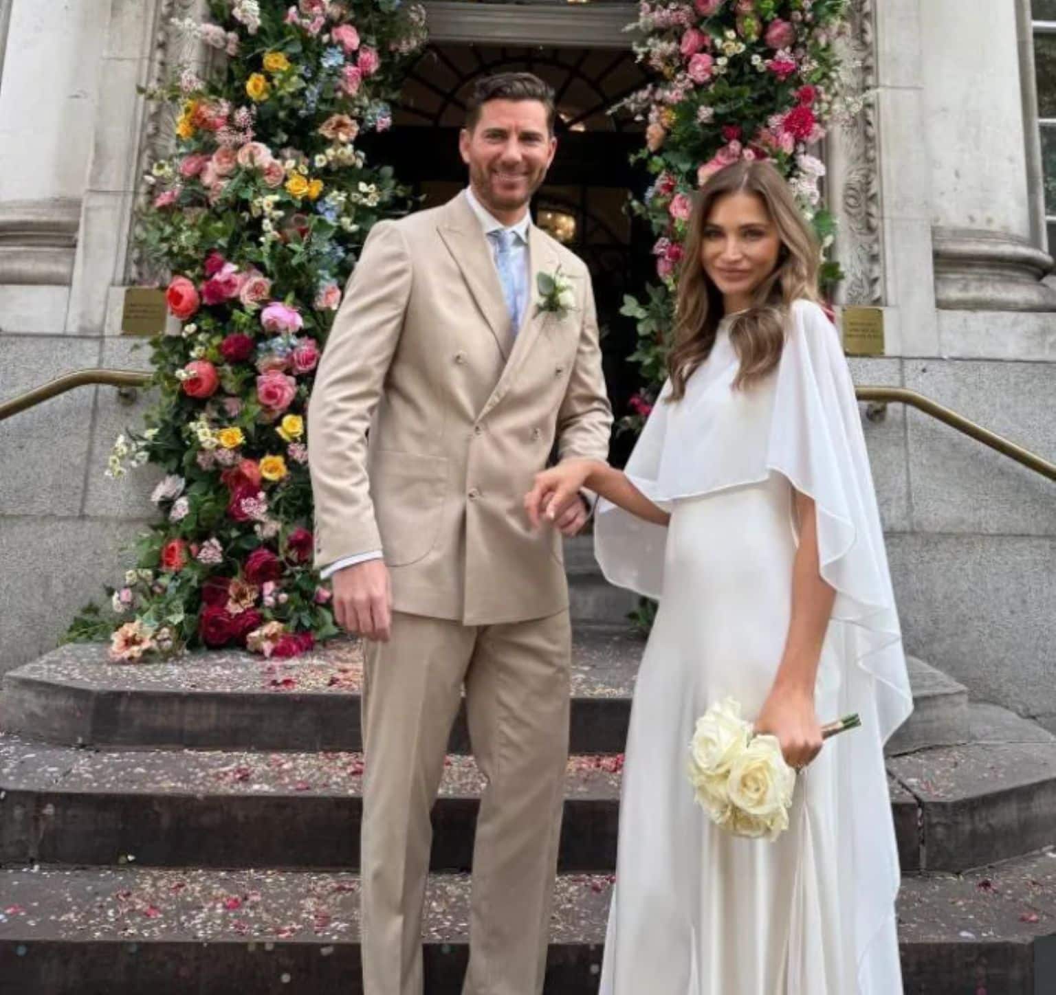 Couple de mariés posant sur les marches d’un bâtiment orné d’une arche de fleurs, la mariée porte une robe fluide blanche avec cape en tulle et un bouquet de roses ivoire.