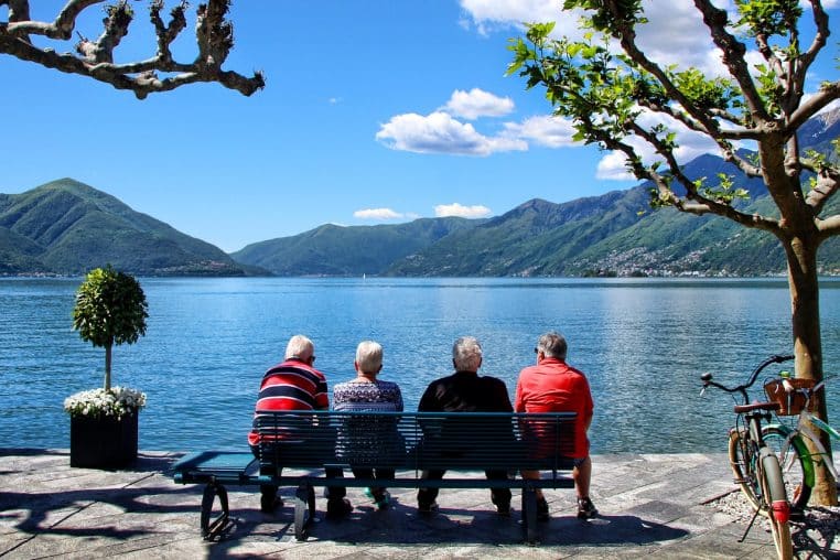 Deux personnes âgées de dos, assises sur un banc face à un lac, paysage calme en format horizontal.