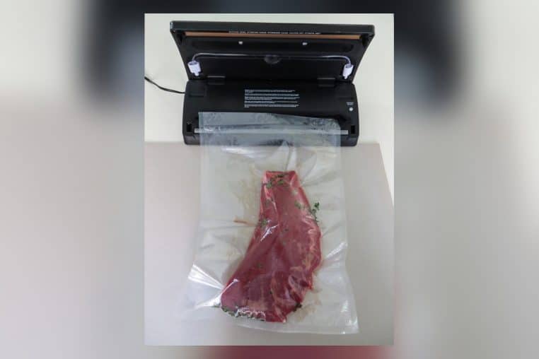Appareil de mise sous vide ouvert avec un grand sachet contenant un morceau de viande et des herbes.