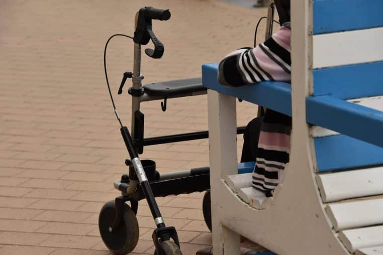 Homme âgé avec déambulateur assis sur un banc extérieur, posture de repos, cadrage horizontal en journée.