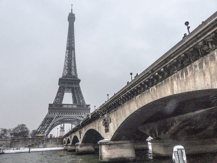 Vue de la tour Eiffel un jour de neige depuis un pont parisien, ciel gris, berges et structures légèrement blanchies, ambiance de froid humide en bord de Seine.