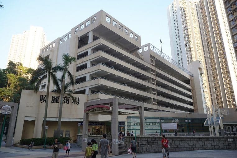 Façade de centre commercial avec parking à étages, palmiers et familles marchant devant l’entrée principale animée.