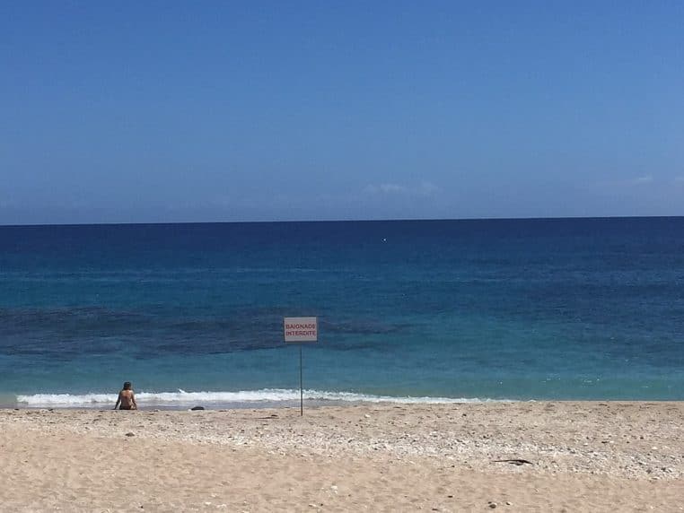 Panneau baignade interdite planté sur le sable de Boucan Canot, face à l’océan calme et à une baigneuse assise au bord de l’eau
