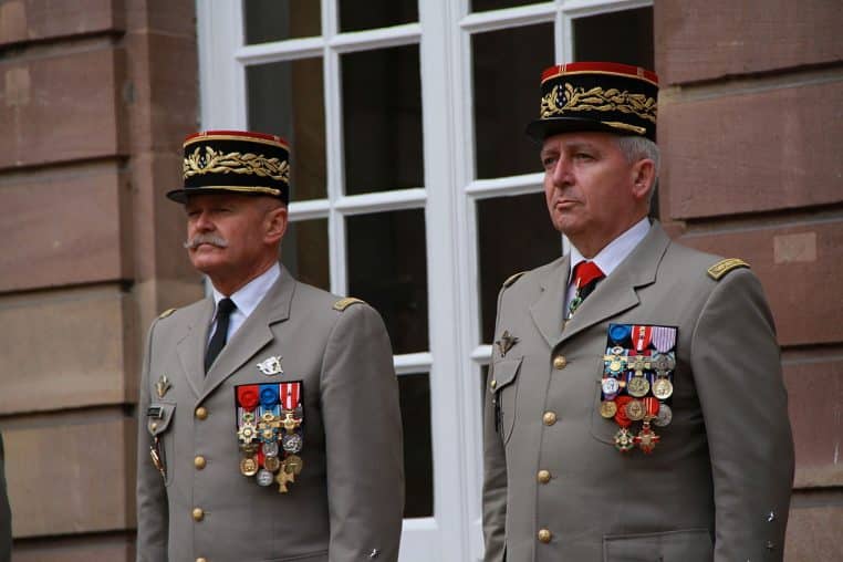 Deux généraux français en uniforme brodé et médaillés se tiennent au garde-à-vous devant une façade en pierre rosée.
