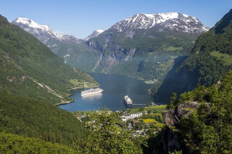 Un paquebot traverse le Geirangerfjord en Norvège, vu depuis la berge, sur une mer calme entourée de montagnes verdoyantes et de falaises abruptes.