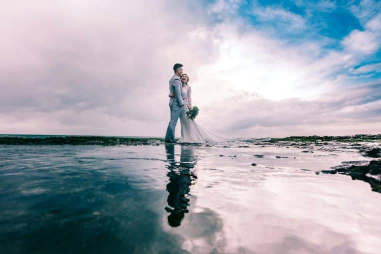 A joyful couple embracing on the beach in wedding attire under a dramatic sky.