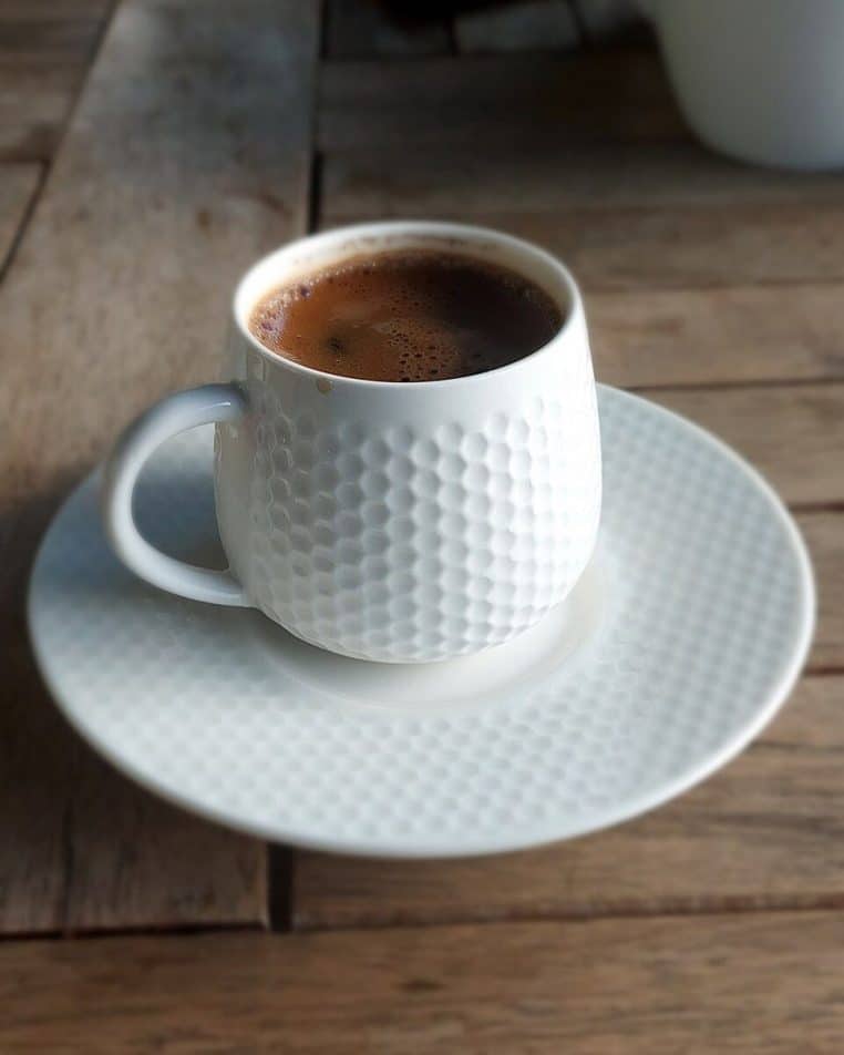 Tasse de café turc dans une coupelle blanche, remplie d’un café sombre mousseux, posée sur une table claire en intérieur.
