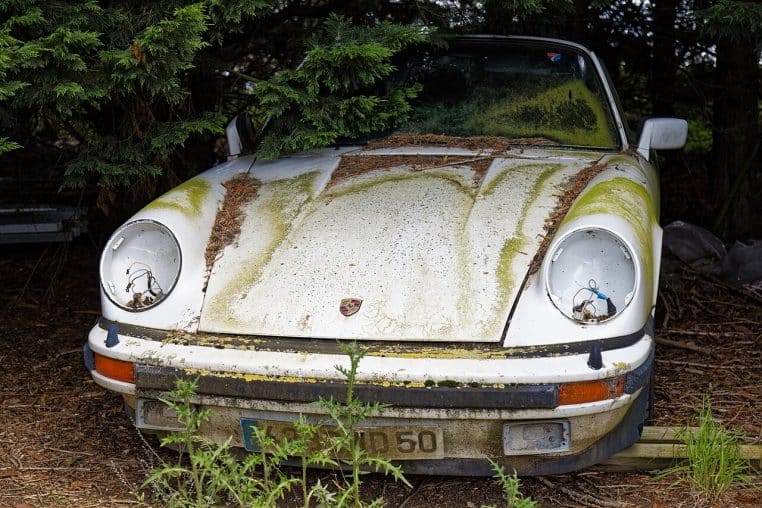 Vieille Porsche abandonnée en plein air, carrosserie marquée par la rouille et la saleté après des années d’exposition aux intempéries.
