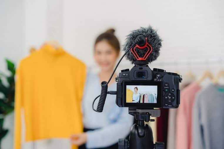 Jeune blogueuse mode asiatique filmant une vidéo de présentation de vêtements avec un appareil photo sur trépied dans son salon lumineux