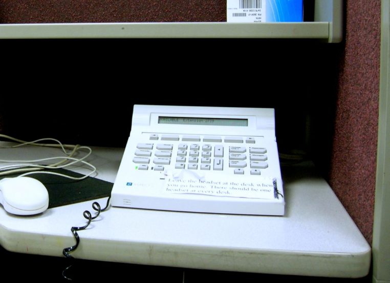 Téléphone de poste dédié à un call center posé sur un bureau, entouré de câbles et d’accessoires informatiques, symbole du démarchage intensif.