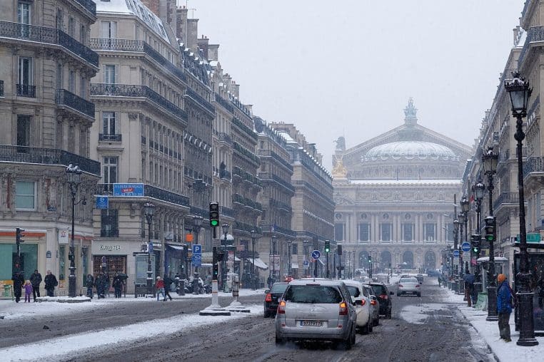 Avenue parisienne menant à l’Opéra Garnier, chaussée enneigée, voitures et piétons dans une lumière grise, façades haussmanniennes soulignant l’ambiance hivernale.