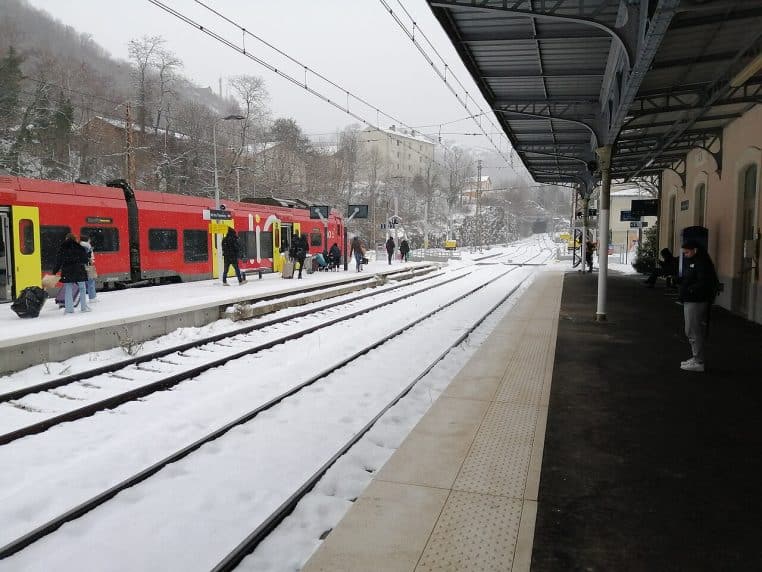 Quai de la gare d’Ax-les-Thermes sous la neige avec un TER rouge et jaune à quai, voyageurs et rails recouverts d’une fine couche blanche.