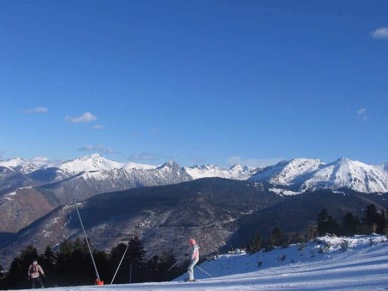 Panorama horizontal d’Ax-Bonascre avec chalets et sapins enneigés, ciel d’hiver voilé, pente douce descendant vers le front de neige.