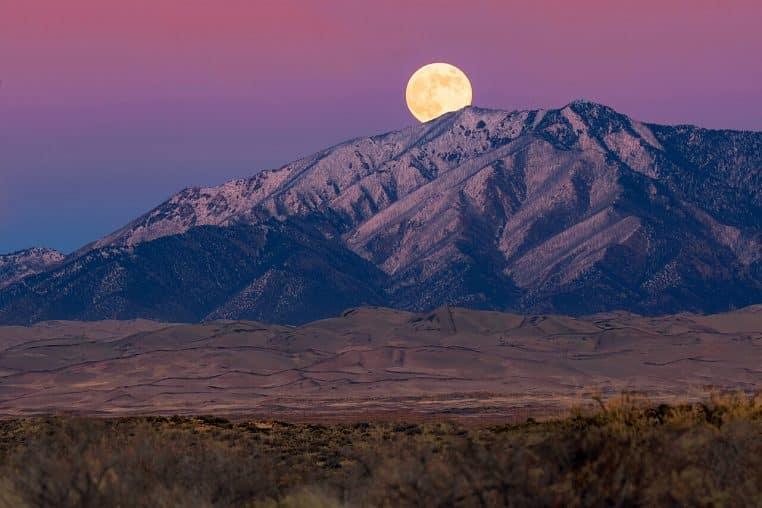 Lune du Castor dorée se levant au-dessus de dunes et d’un sommet enneigé au crépuscule, ciel bleu profond, ambiance automnale.