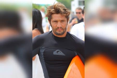 Bixente Lizarazu en combinaison de surf noire tient une planche orange, le regard concentré, devant une foule floue.