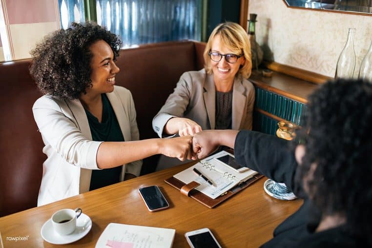 Poings entrechoqués de deux collaboratrices souriantes dans un café, signe d’accord après échange, cadrage horizontal.