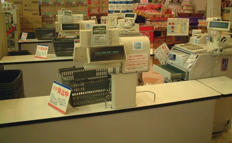 Rangée de caisses enregistreuses vides dans un supermarché japonais, avec paniers noirs alignés et signalétique en japonais.