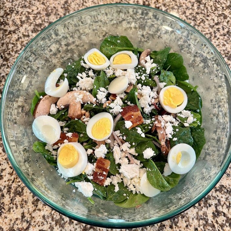 Salade généreuse d’épinards avec œufs, fromage émietté et morceaux de bacon dans un grand saladier transparent sur un plan de travail.