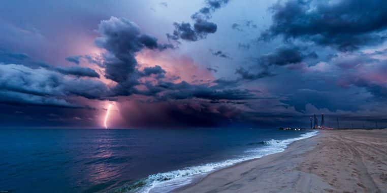 Plage déserte au crépuscule, ciel chargé de nuages sombres et éclair violet tombant en mer, vagues calmes s’écrasant sur le rivage.