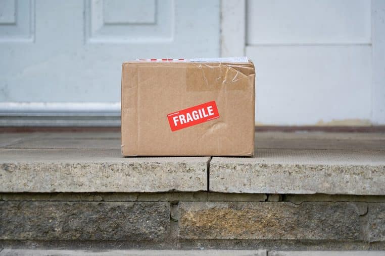 Colis en carton avec étiquette rouge « Fragile » posé devant une porte, angle du carton froissé qui évoque un transport raté.