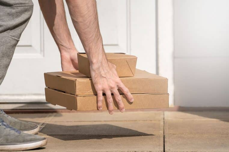 Mains d’un livreur déposant trois boîtes en carton empilées devant une porte d’entrée blanche, prêtes à être récupérées par le client.